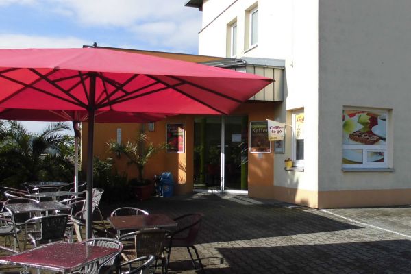 Café Haideblick der Bäckerei und Konditorei Zum Kirchbäck Burgstädt Haus mit Anbau, in dem sich das Café Haideblick befindet. Davor roter Sonnenschirm mit Stühlen und Tischen darunter.