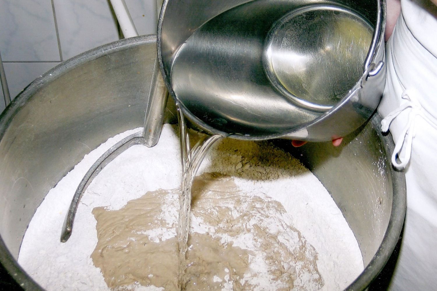 Mit reichlich Futter aus Mehl und Wasser zieht der Sauerteig in einen Kneter um und ruht wieder. Ein Bäcker gibt Mehl und Wasser in einen Kneter.