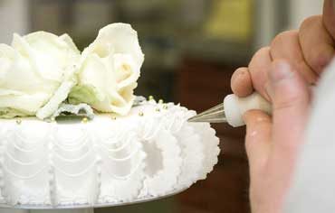 Frau dekoriert mit Spritztüte eine weiße Torte mit zwei weißen Rosen darauf